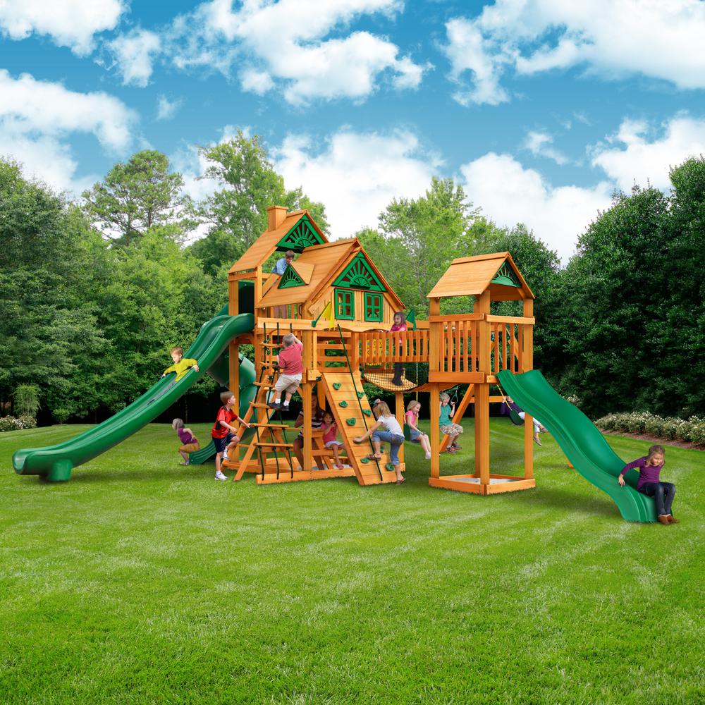 Wooden playset with two green slides, climbing walls, rope ladder, swings, and children playing on grass outdoors