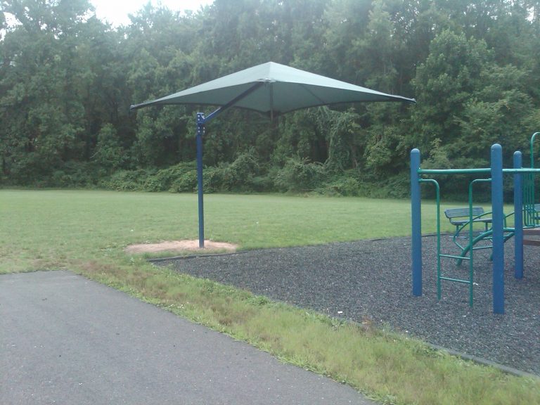 Cantilever square umbrella with side post and taut fabric providing shaded area over green grass and playground trees