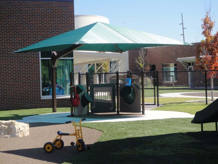 Green cantilever square shade umbrella with single side post over playground area featuring play panels and yellow tricycle