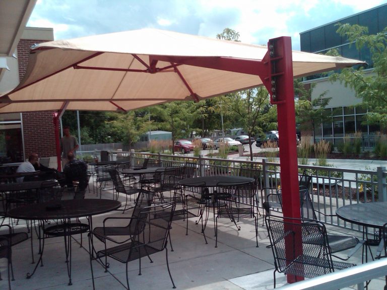 Outdoor patio with black metal tables and chairs shaded by a large beige cantilever square umbrella with a red frame