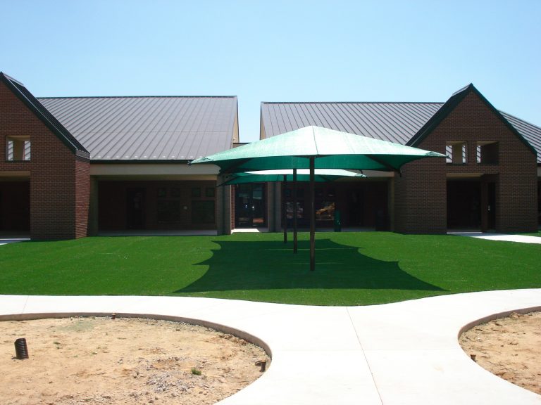 Green square center-post umbrella shade canopy providing UV protection over artificial grass in an outdoor courtyard setting