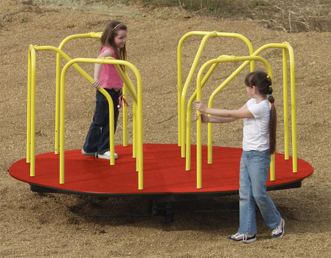 Red powder-coated steel merry go round with yellow handrails on playground surface, two girls holding bars playing