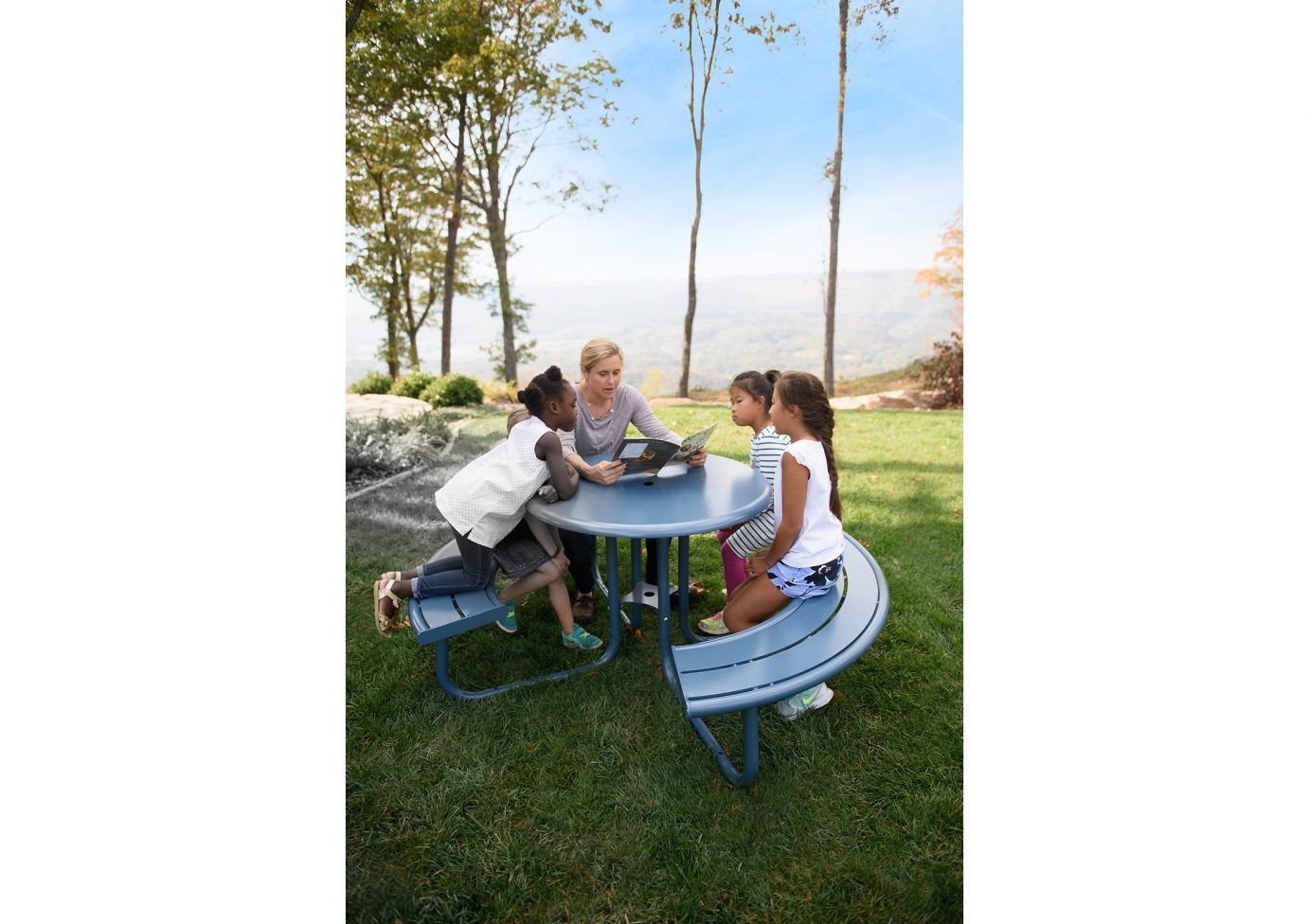 Blue round outdoor picnic table with curved benches, children and adult reading, grassy park setting