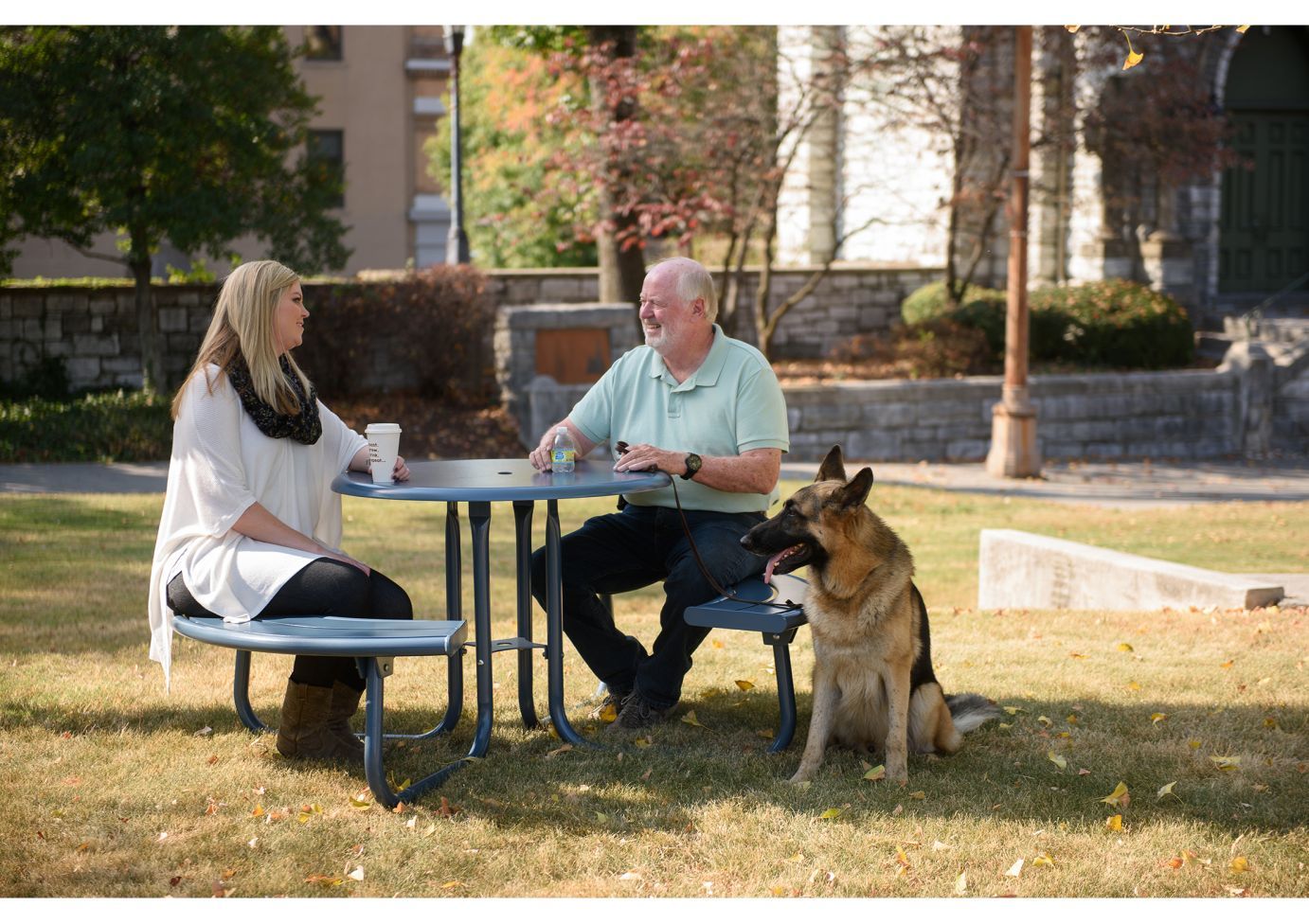 Round metal picnic table with attached benches, two people seated, German Shepherd dog on grass, outdoor setting.