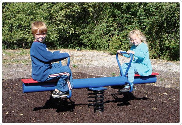 Two children on a blue and red spring rider seesaw with handles on a playground surface with mulch and greenery behind