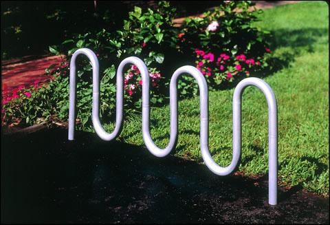 White powder-coated tubular steel bike rack with seven wavy loops installed on black pavement near green grass and flowers