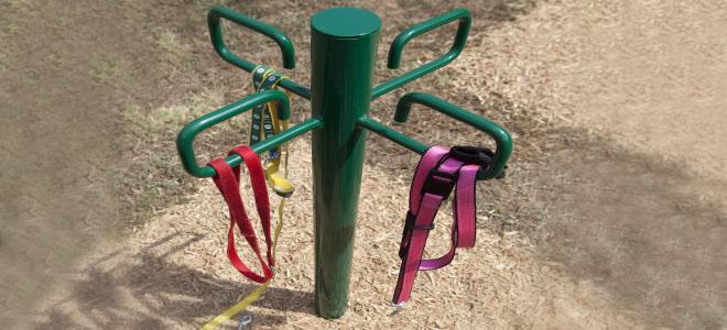 Green metal leash post with four C-hooks holding colorful dog leashes on dirt ground at a dog park