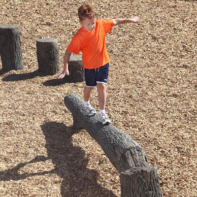 Child balancing on Nature Themed Fallen Tree Balance Beam with wood chip ground and tree stump stepping posts in outdoor playground