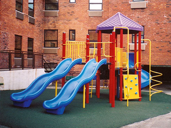 Colorful modular playground with multiple blue slides, spiral climber, rock wall, and yellow railings on green safety surface