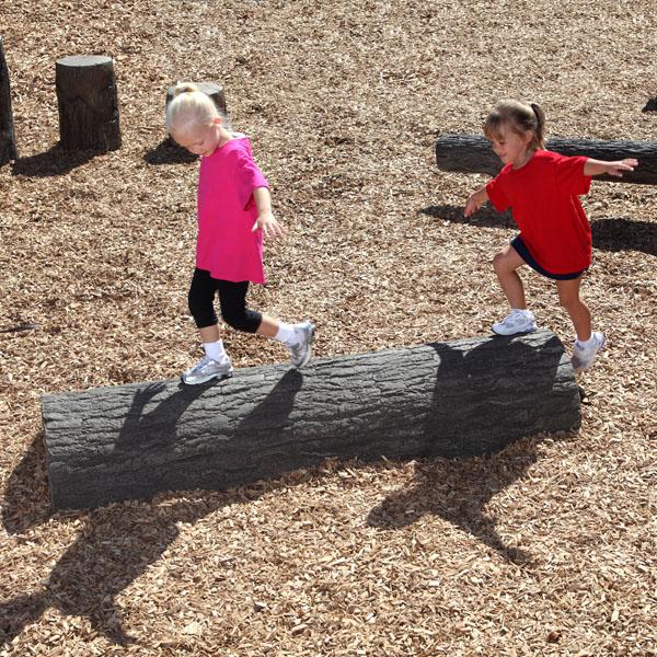 Children playing on a nature-themed half log balance beam in an outdoor setting