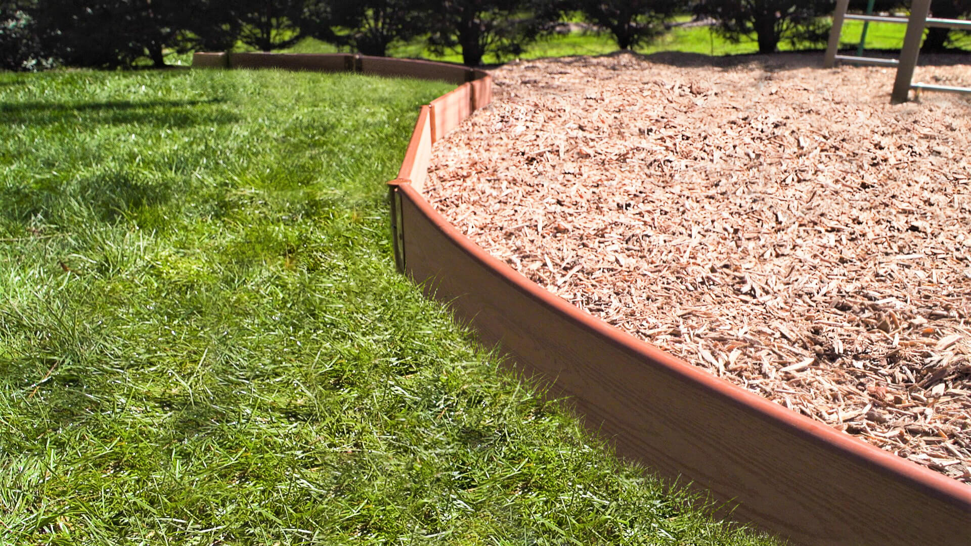 Tool-Free Classic Curved Playground Border in sienna containing wood chip safety surfacing, bordered by green grass and trees.