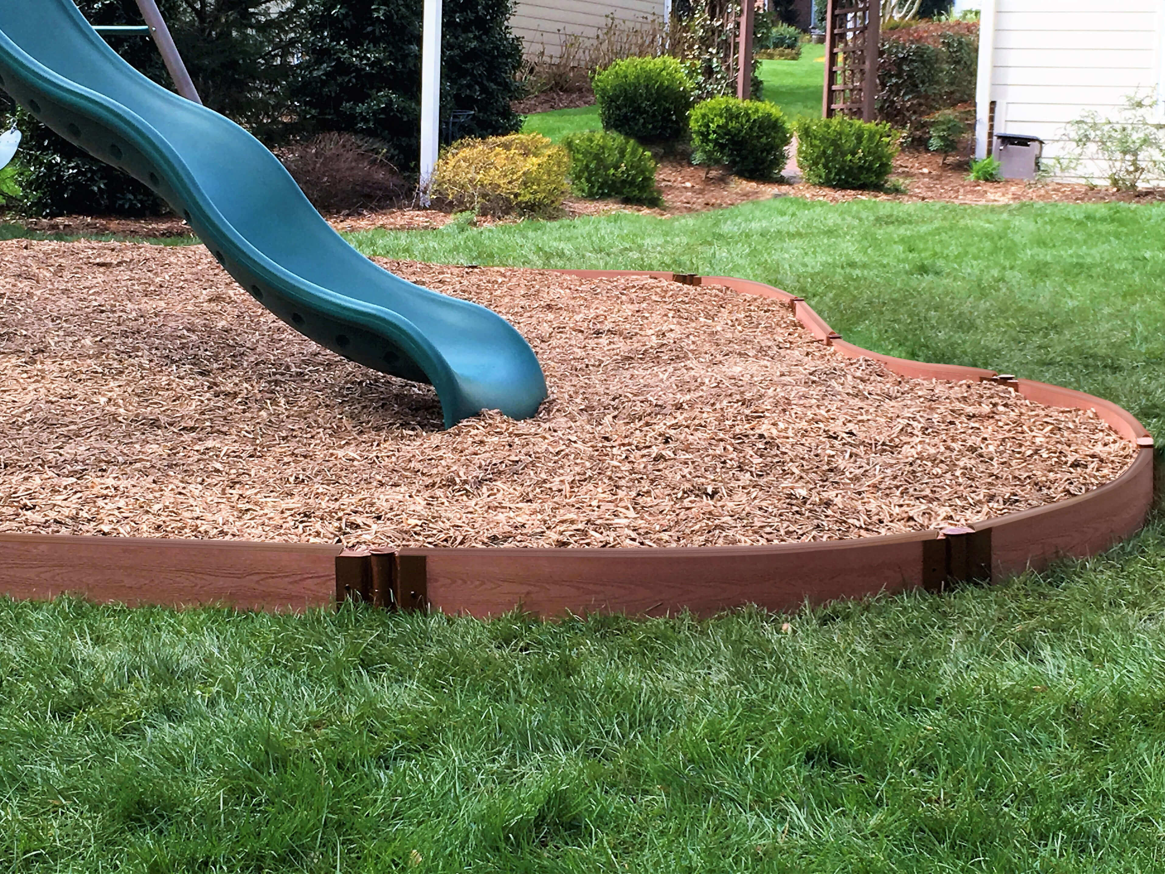 Curved brown playground border made of recycled plastic and wood fibers containing wood mulch around a blue slide in a backyard setting