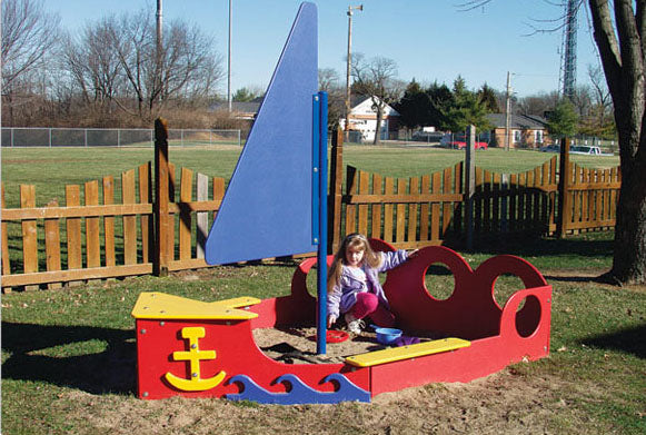 Colorful sailboat-shaped sandbox with blue sail and red-yellow accents, child playing inside on grass outdoors
