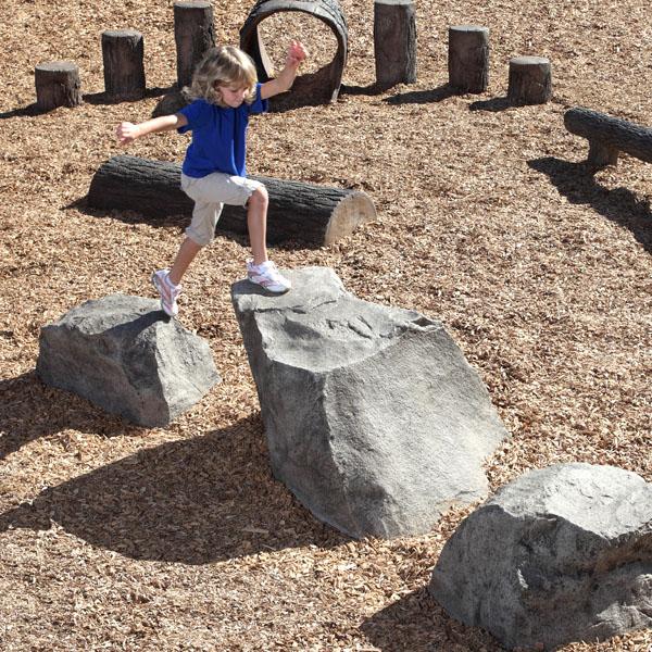Child jumping across three hand-painted Nature Rocks Stepping Boulders in a woodchip playground with climbing and balance features
