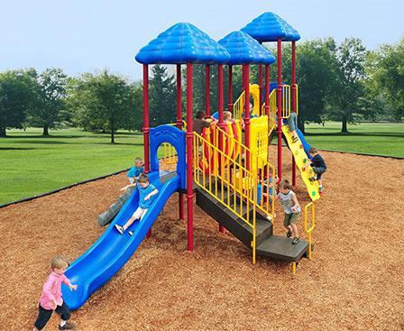 Kids playing on commercial playground with three blue shade roofs, slides, climbers, and a clatter bridge in a mulched park.