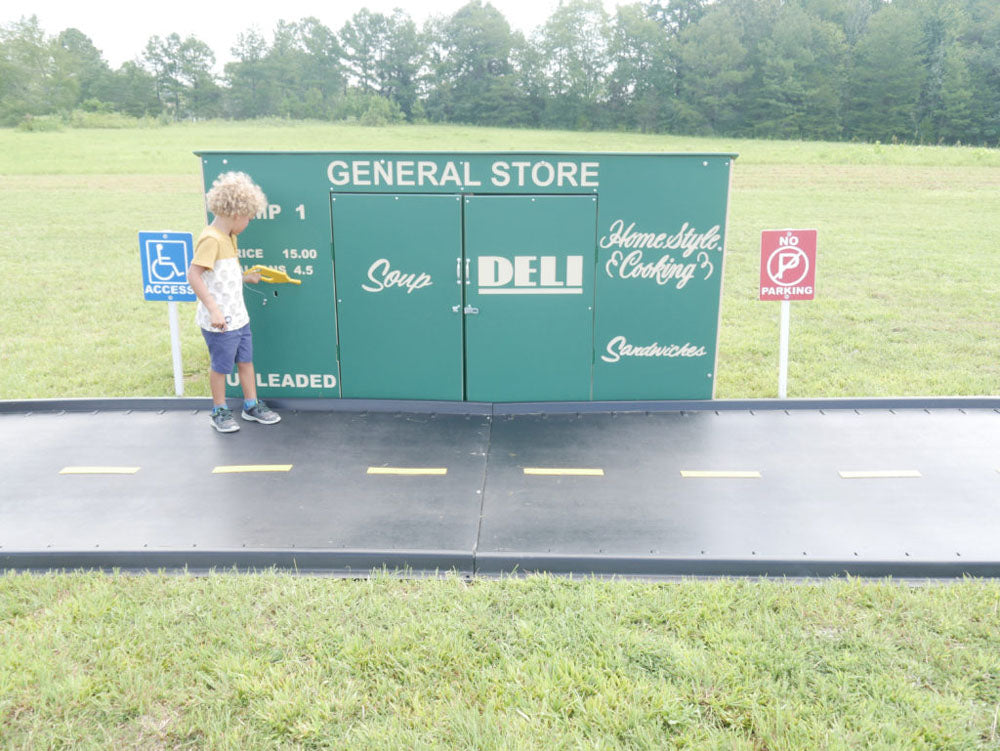 Child playing at a green outdoor playhouse designed as a general store with trike paths and accessibility signs.