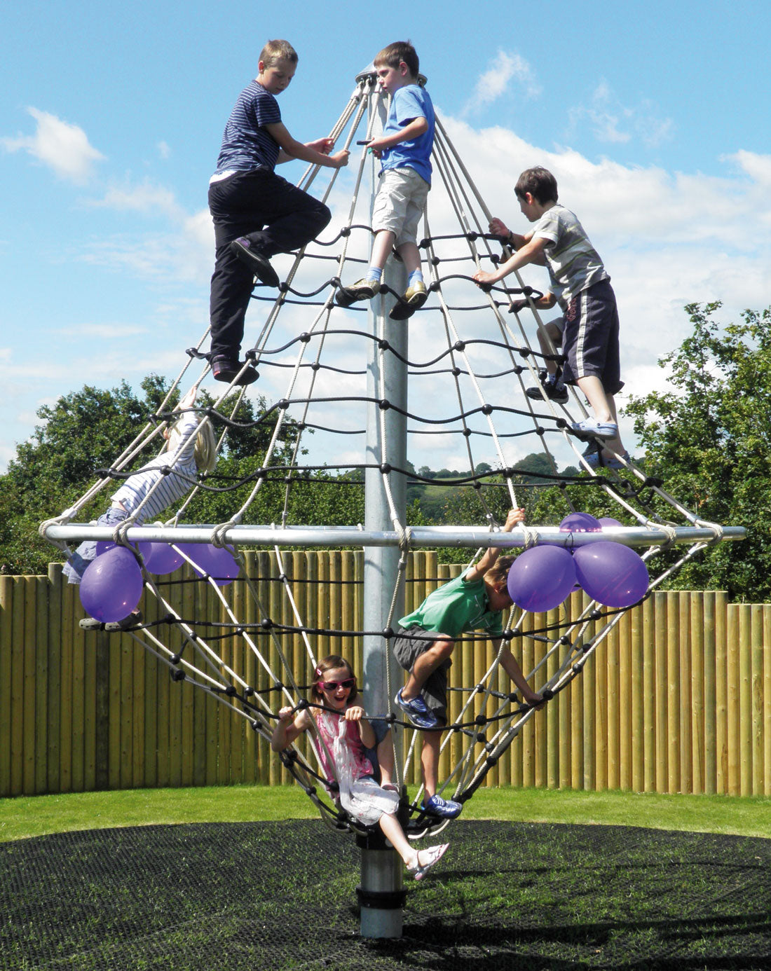 Children playing on a rope climbing structure with a central metal pole and purple balloons outdoors