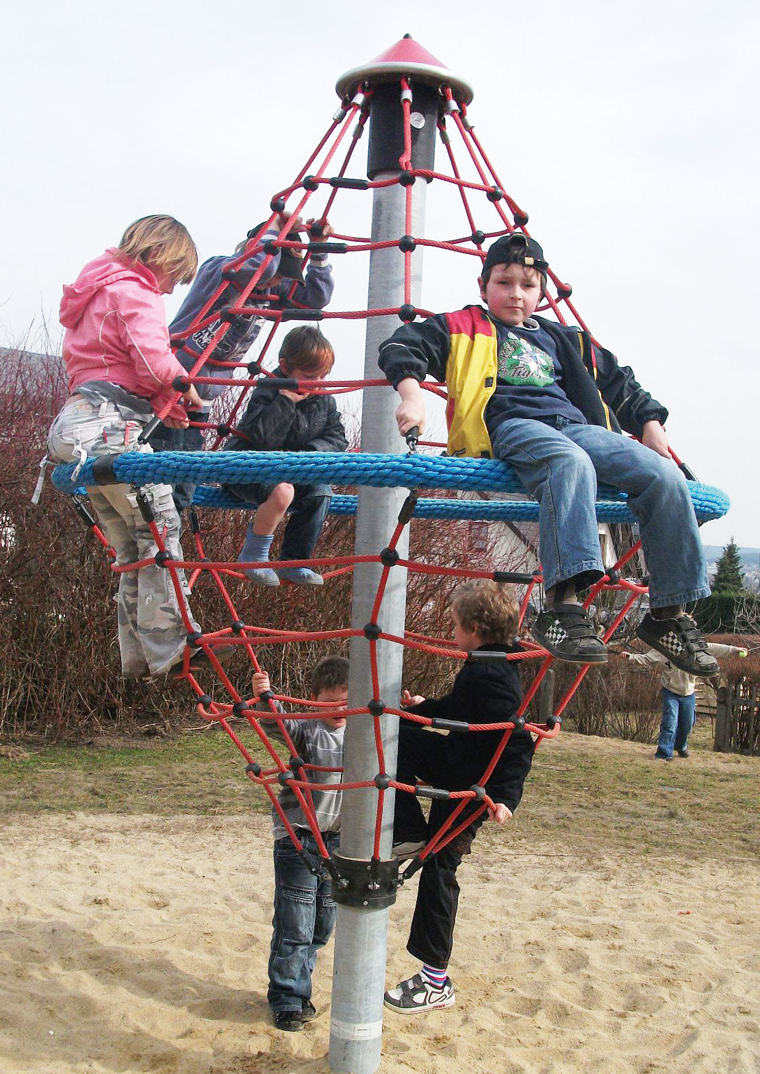 Children climbing and sitting on a red rope climbing structure with a blue circular net on a sandy playground
