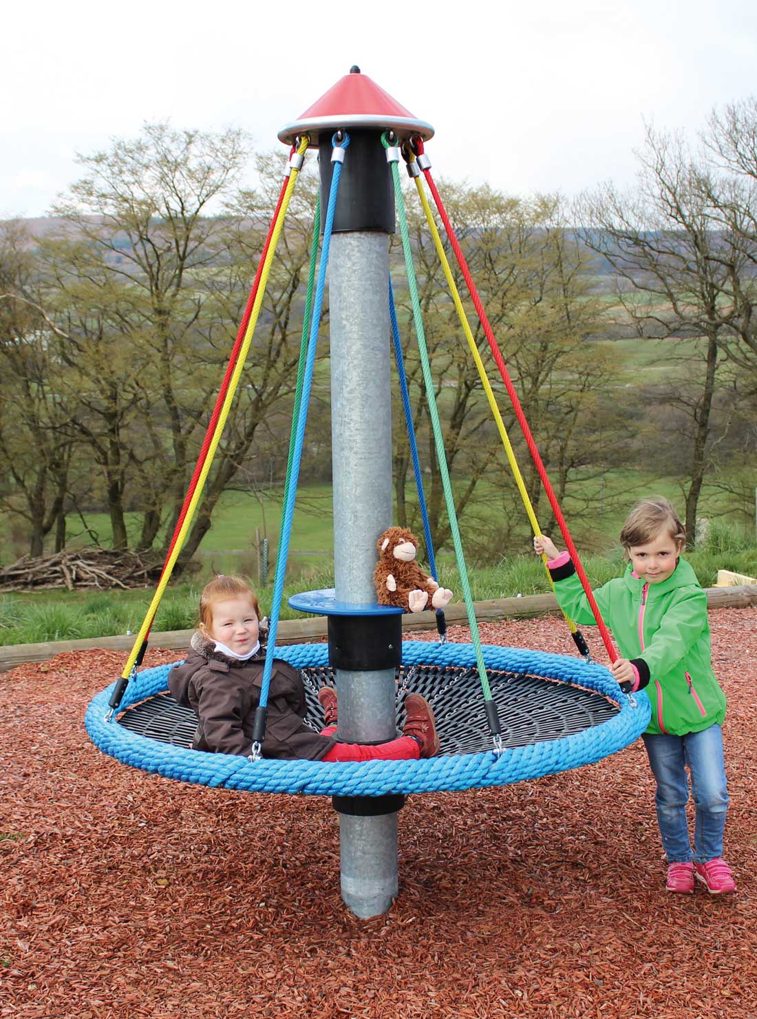 Child sitting inside blue rope nest seat of playground carousel, suspended by colorful ropes, another child pushing