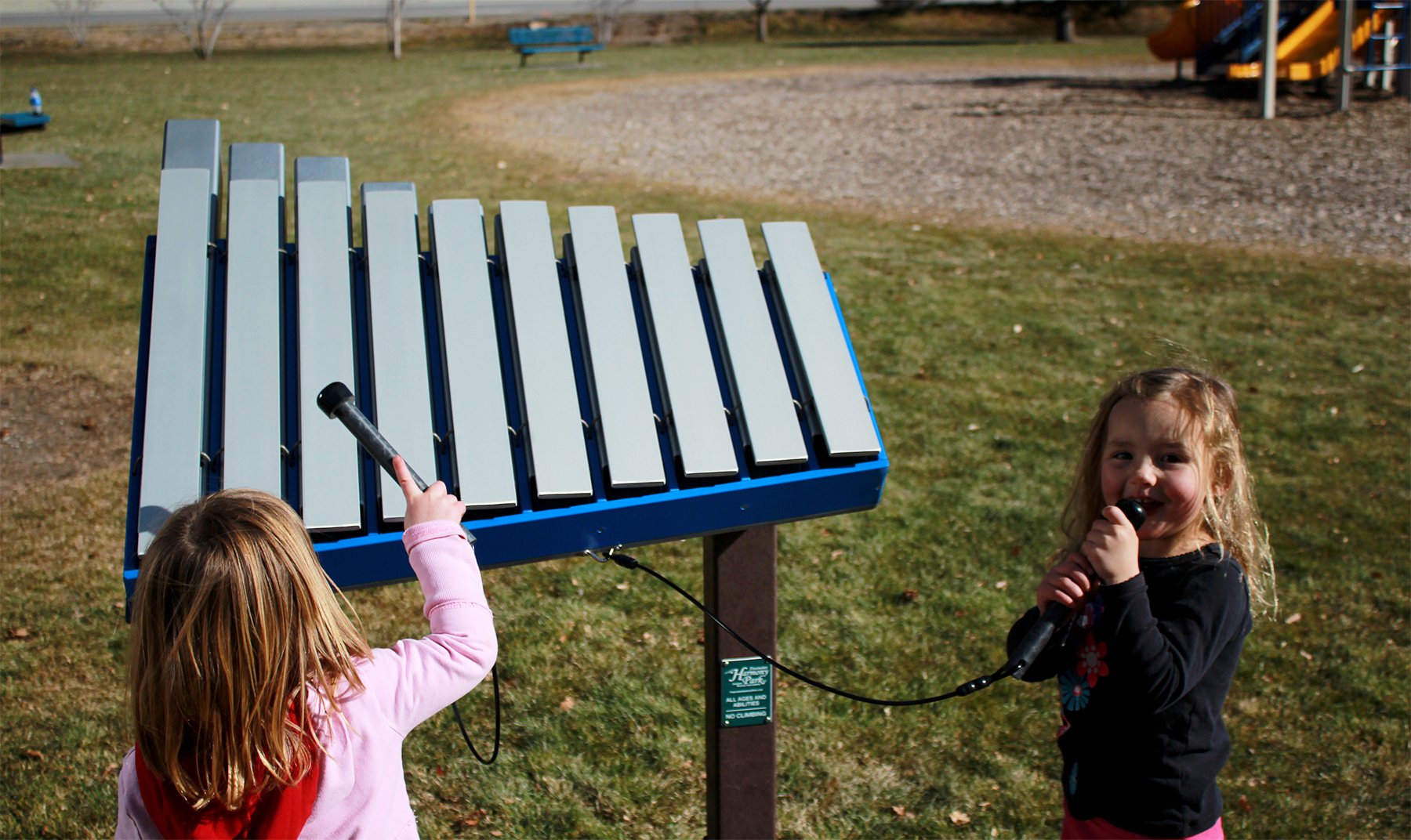 Outdoor blue aluminum bar musical instrument with resonating tubes played by two children in a park setting