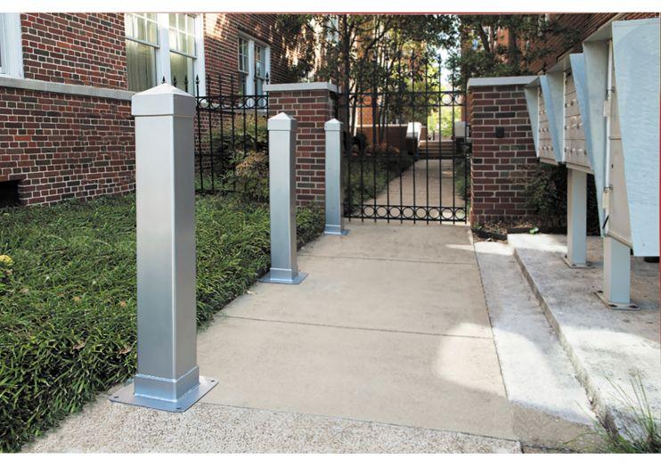 Three powder-coated steel bollards installed on a sidewalk near a brick building and metal gate