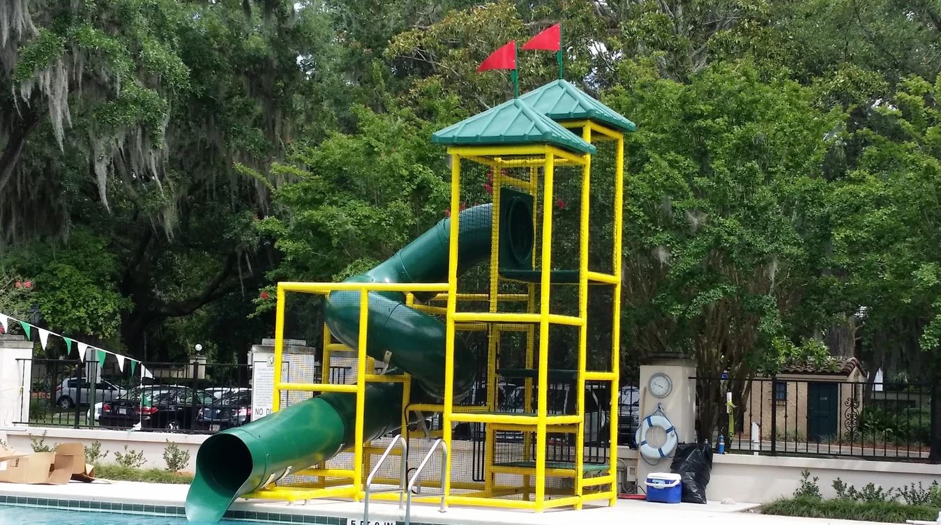 Green spiral water slide with yellow safety netting and two peaked roofs topped with red flags beside a pool