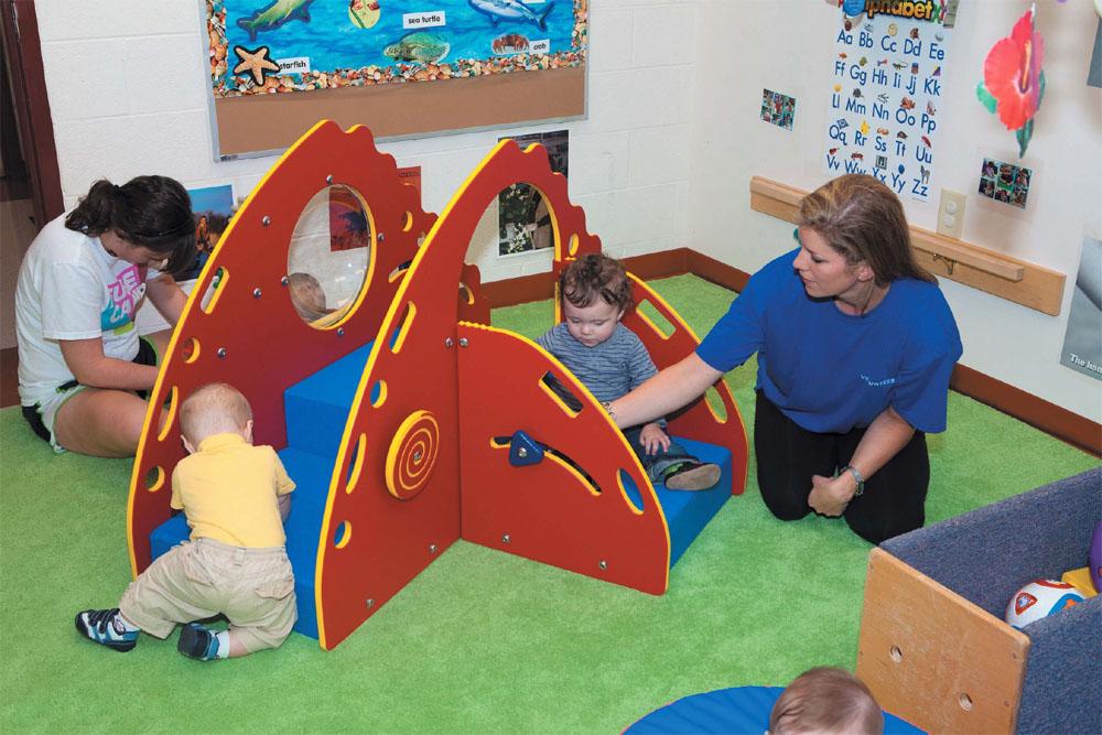 Toddler play area with red and blue soft climbing structure, children crawling and sitting, adult supervising on green carpet
