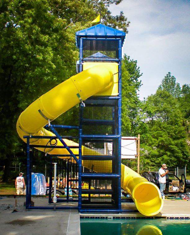 Tall blue-framed water slide with bright yellow enclosed flume curving into a pool, surrounded by trees and outdoor setting