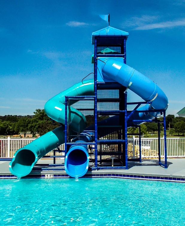 Two curved blue enclosed tube water slides descending from a tall metal frame into a pool with safety netting and a blue roofed platform