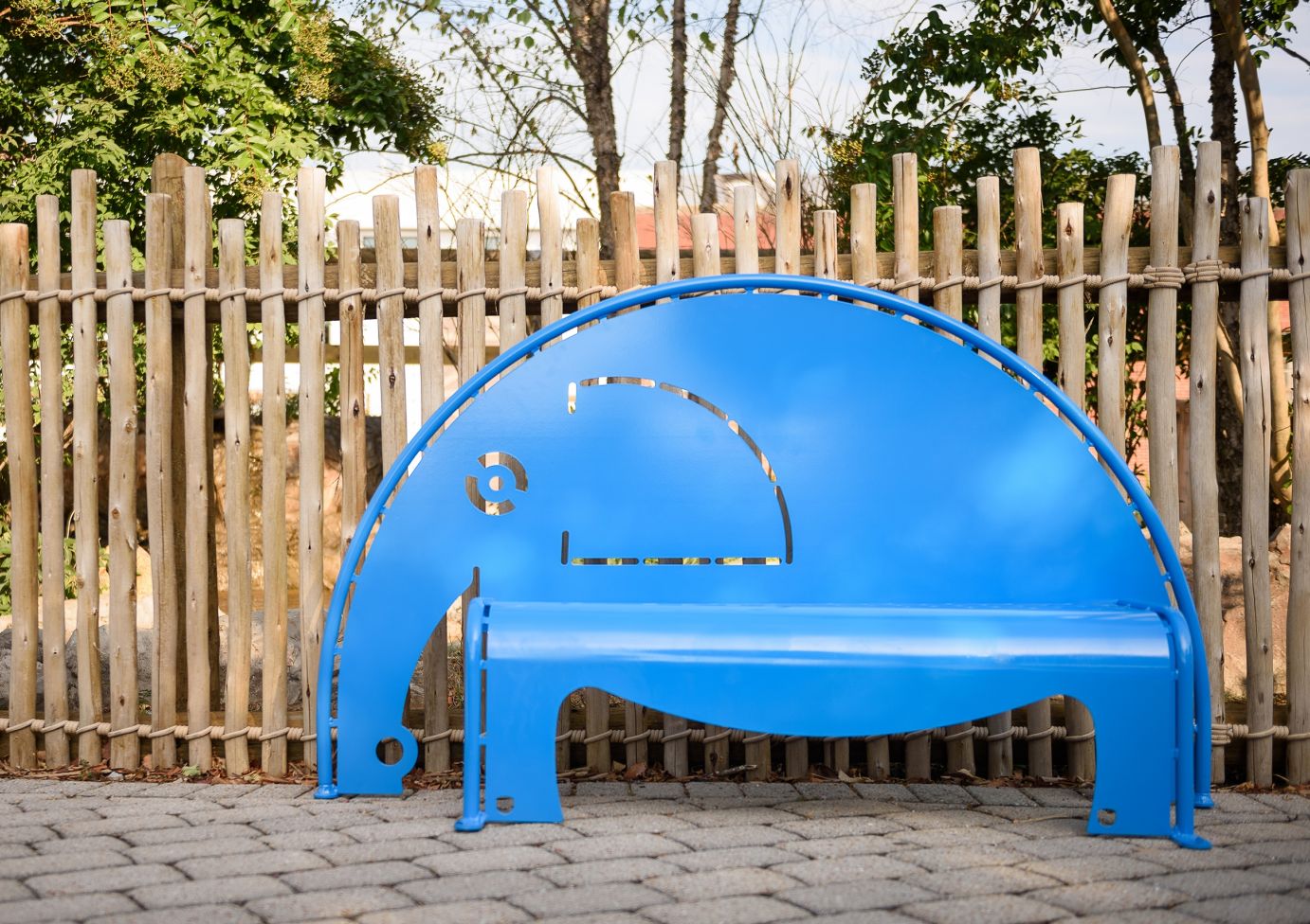 Bright blue elephant-shaped metal bench with cutout details on stone pavement in an outdoor natural setting