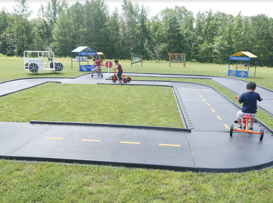 Winding outdoor trike path with black asphalt, yellow lane markings, play buildings, and children riding tricycles on grass