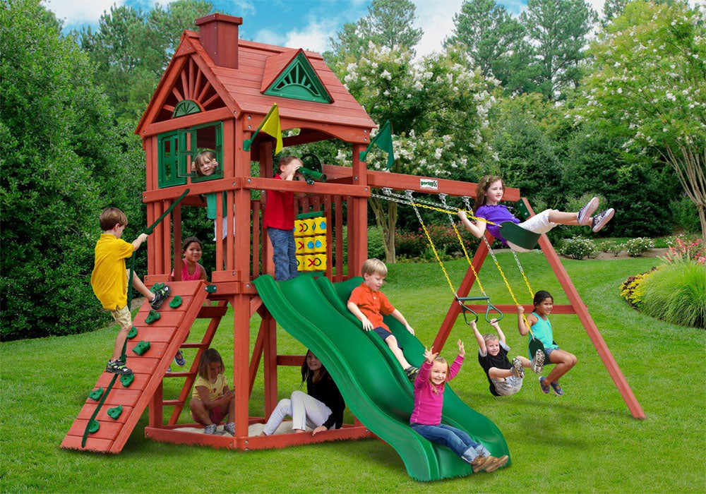 Double Down Wooden Swing Set with two green Alpine Wave slides, rock wall, clubhouse with shutters, swings, and tic-tac-toe panel in backyard.