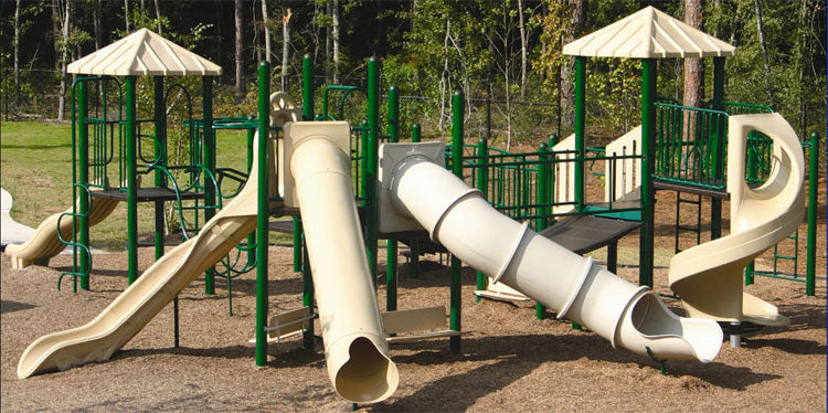 Outdoor modular playground with beige slides, green climbing structures, two pyramid-roofed platforms, and wood chip ground