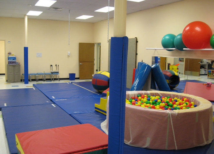 Blue wrap-around safety pole pads on round poles in an indoor playroom with mats, therapy balls, and a ball pit.