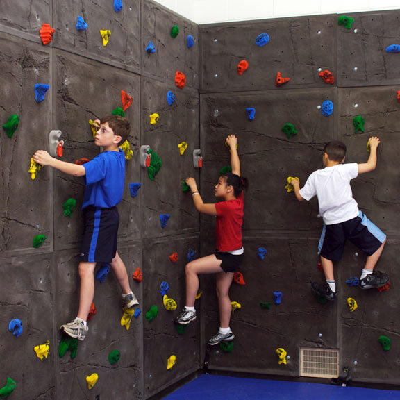 Superior Traverse Climbing Wall Panel with textured gray rock-like surface, multicolor holds, blue mat, and three children climbing indoors
