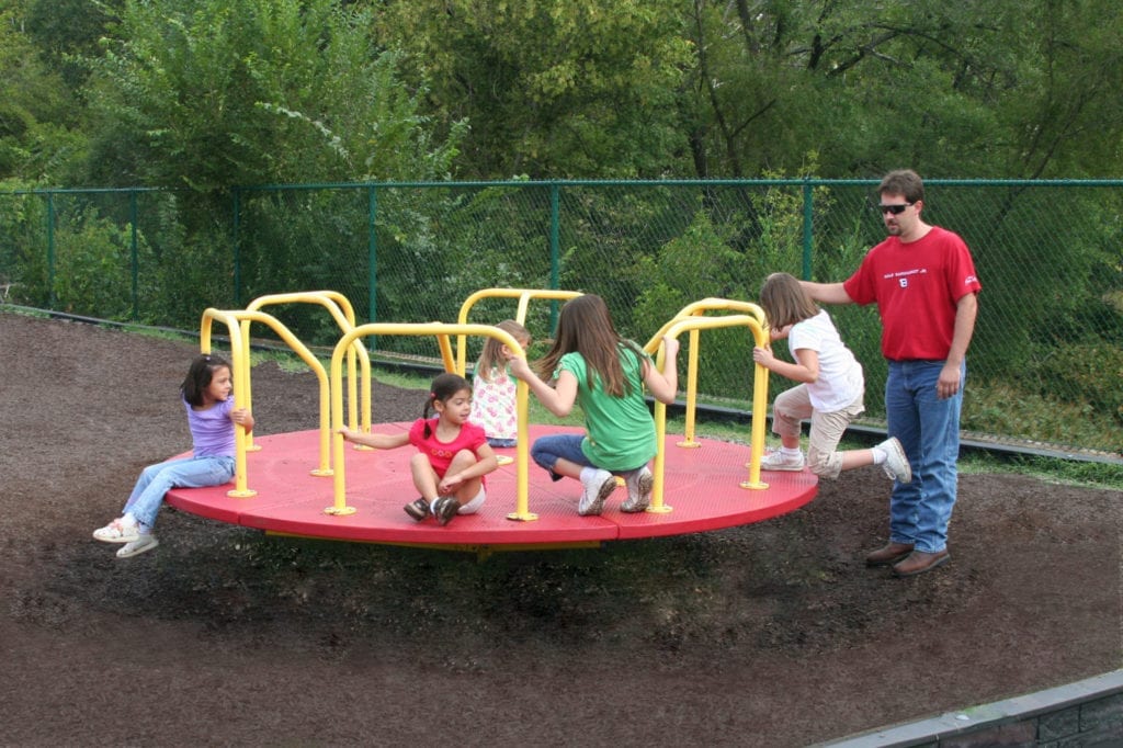 Red merry go round with yellow handrails, children playing, adult supervising, playground mulch surface