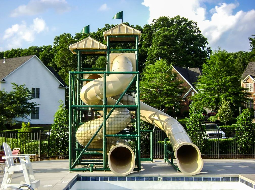 Two beige enclosed water slide flumes with green supports and flags, empty pool in suburban aquatic setting