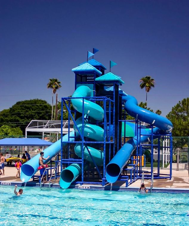 Large blue water slide structure with three twisting enclosed flumes and two flag-topped towers at a public pool