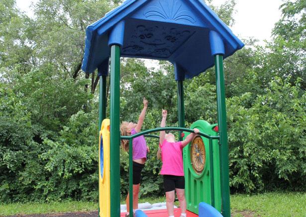 Early childhood playground structure with blue roof, green and yellow panels, dinosaur fossil climber, and two children playing outdoors.