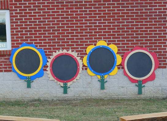 Four colorful flower-shaped outdoor chalkboards with black writing surfaces and green stems mounted on a brick wall