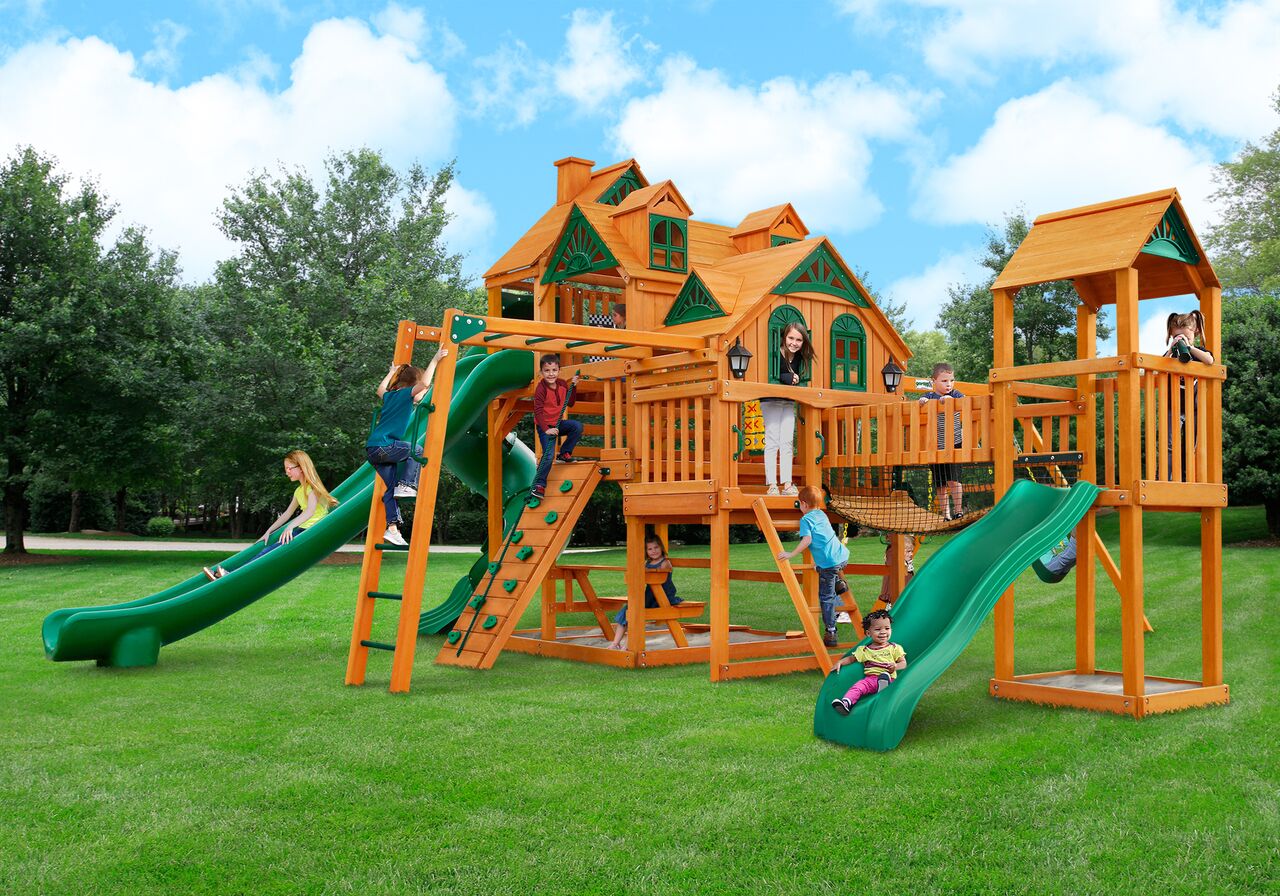 Wooden playset with two slides, climbing wall, monkey bars, tic-tac-toe board, and children playing on grass outdoors