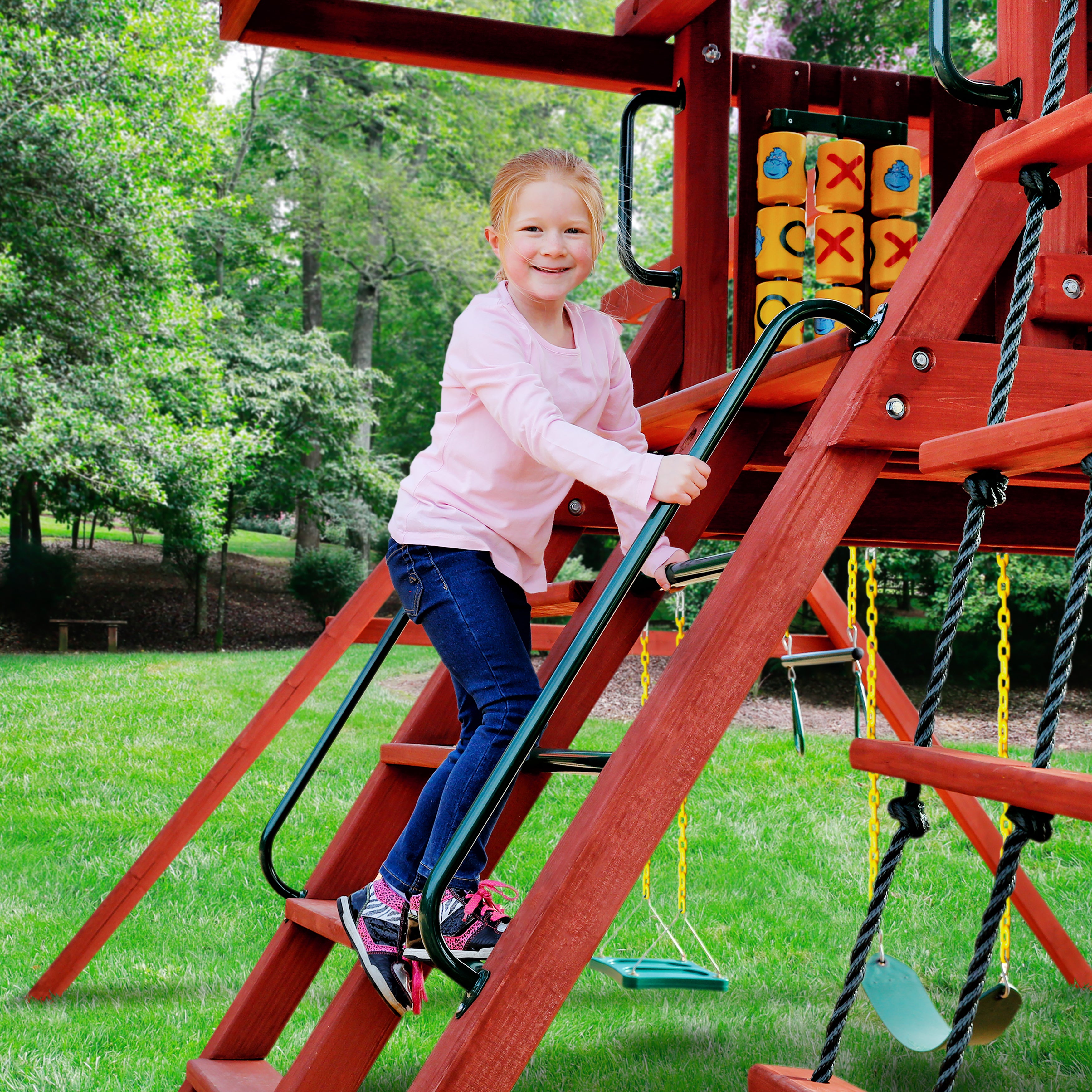 Child climbing wooden playset ladder using green powder-coated metal safety handrails with tic-tac-toe panel and swings nearby