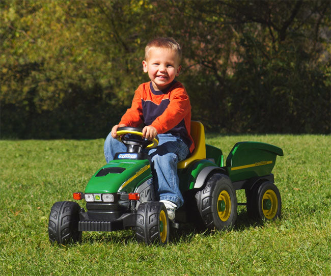 Child riding green pedal tractor with yellow seat and matching detachable trailer on grass outdoors
