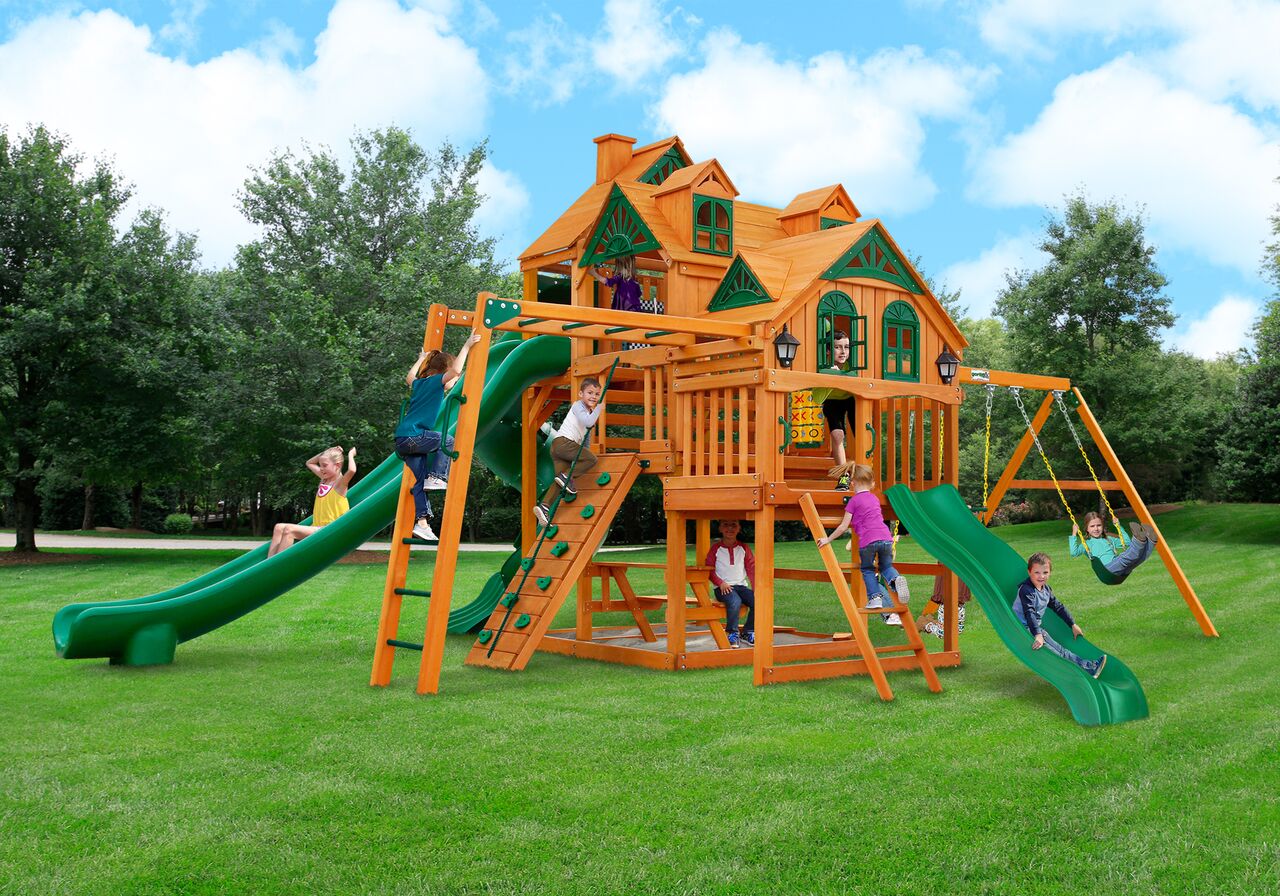 Cedar wooden playset with three green slides, climbing wall, monkey bars, swings, and playhouse features with children playing