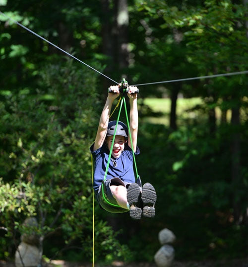 Child wearing helmet on green disc seat using Alien Flier X3 Zip Line Base Kit trolley through wooded outdoor area