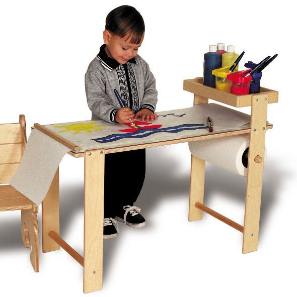 Child painting at a sturdy wooden art table with paper roll, paint containers, and brushes on side storage tray