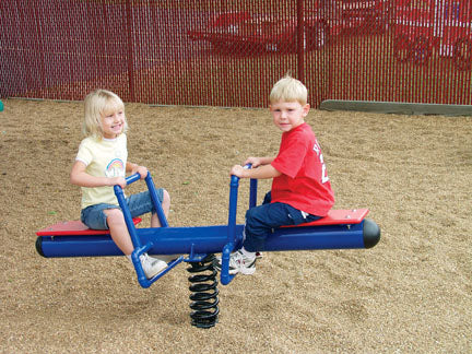 Teeter Spring Rider with two children playing on blue and red spring-based see-saw in playground with wood chip ground and red fence background