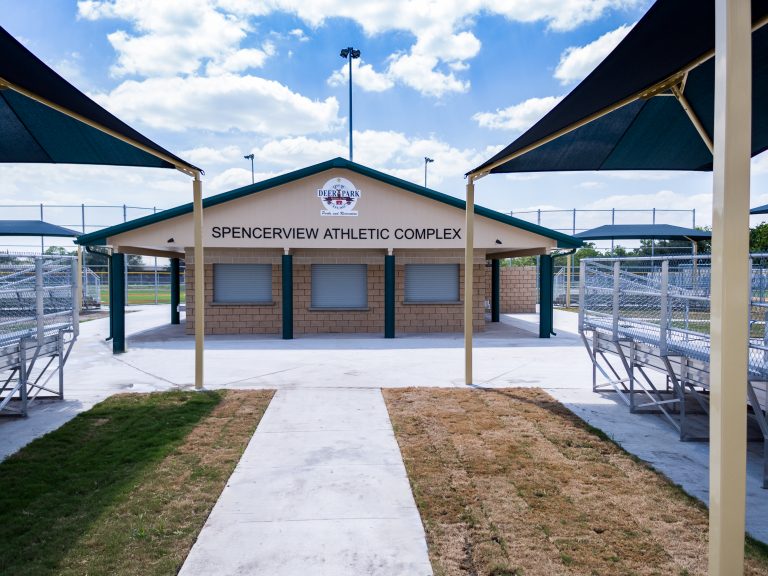 Rectangular hip roof shade structure with four posts and dark fabric covering bleacher seating at athletic complex