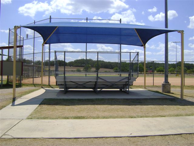 Rectangular blue hip roof shade structure with four posts covering metal bleachers at a baseball field
