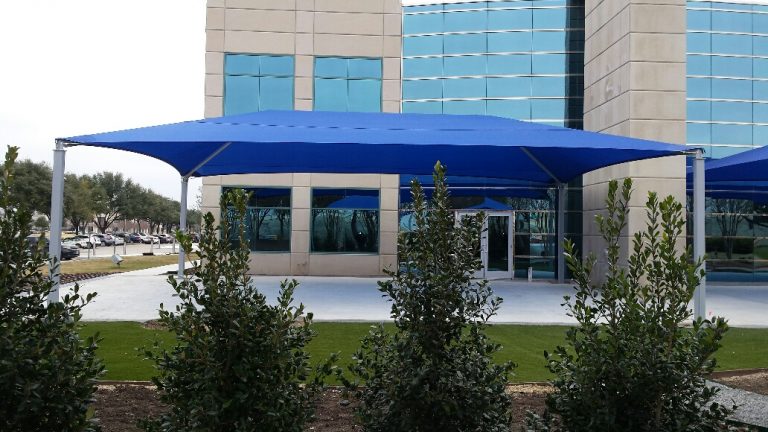 Rectangular blue hip roof shade structure with four metal posts over outdoor area near modern building